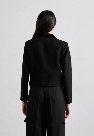 Black cropped jacket made of textured fabric, featuring a classic collar and long sleeves, paired with black trousers.