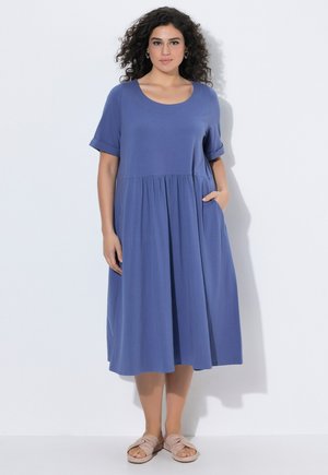 Kvinde med krøllet hår iført en løs, mid-lang blå kjole med lommer og opsmøgede ærmer, stående foran en ensfarvet hvid baggrund.