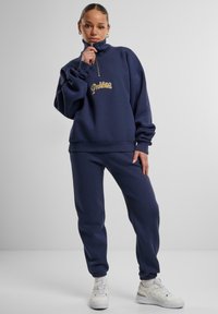 Felpa con zip blu navy con la scritta "Proibito" in oro, abbinata a pantaloni jogger coordinati; realizzata in tessuto morbido, presenta una vestibilità ampia e polsini a costine.