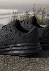 Paire de chaussures de sport noires avec semelles Cloudfoam reposant sur un trottoir avec des plantes floues et le ciel en arrière-plan.