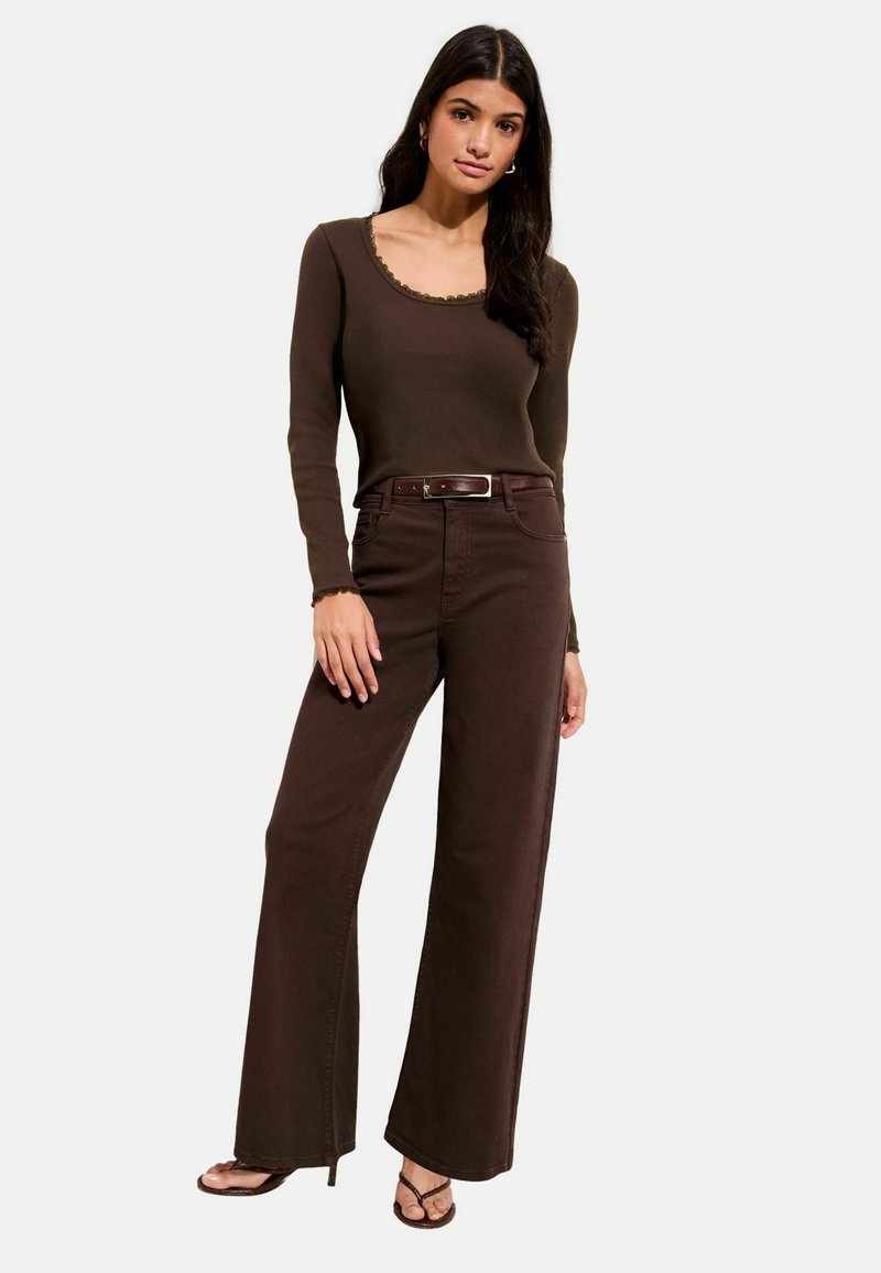 Hnedý top s dlhými rukávmi a vlnitým výstrihom, v kombinácii s hnedými nohavicami s vysokým pásom a širokými nohavicovými nohavicami a čiernymi sandálmi na podpätku.