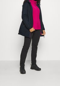 Veste isolante bleu marine avec capuche, portée sur une polaire rose zippée, associée à un pantalon noir et des chaussures de randonnée foncées ; coupe décontractée et design fonctionnel.