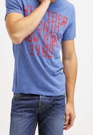 Mężczyzna ubrany w niebieski, nakrapiany t-shirt z czerwonym napisem „Tommy Hilfiger Denim New York 1985” oraz ciemnoniebieskie dżinsy, ręka przy ustach.