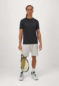 T-shirt noir à manches courtes avec le logo "BOSS", associé à un short gris clair et des baskets blanches. La personne tient une raquette de tennis.