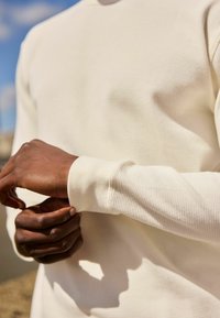 Cremefarvet, struktureret langærmet skjorte med ribbet mønster og tætsiddende manchetter. Hænder justerer ærmet og fremviser stofdetaljer.