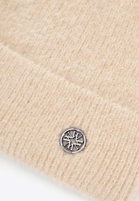 Gorro de punto beige hecho de material suave, con un patrón acanalado texturizado y un emblema redondo de copo de nieve en tono plateado.