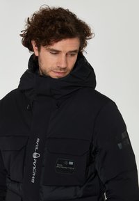 Svart puffjacka med hög krage, stora framfickor och logodetaljer. Slät yta, justerbar huva med dragsnören.