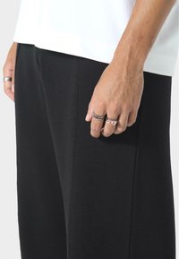 Czarne zwężane spodnie wykonane z miękkiego materiału, charakteryzujące się gładką fakturą i subtelnym połyskiem. Wyeksponowane z białą koszulą i pierścionkami na ręce.