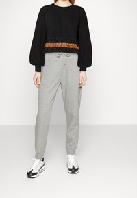 Sweat-shirt noir court avec franges orange texturées et accents à motifs, associé à un pantalon de survêtement gris clair fuselé et des baskets.
