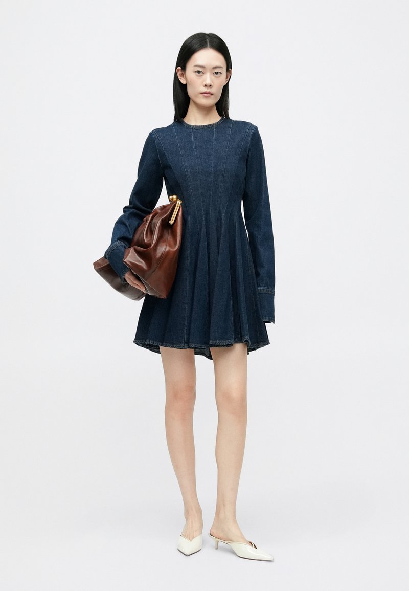 Robe en denim à manches longues, jupe évasée et détails de couture verticaux ; associée à un sac en cuir marron et des mules blanches à bout pointu.