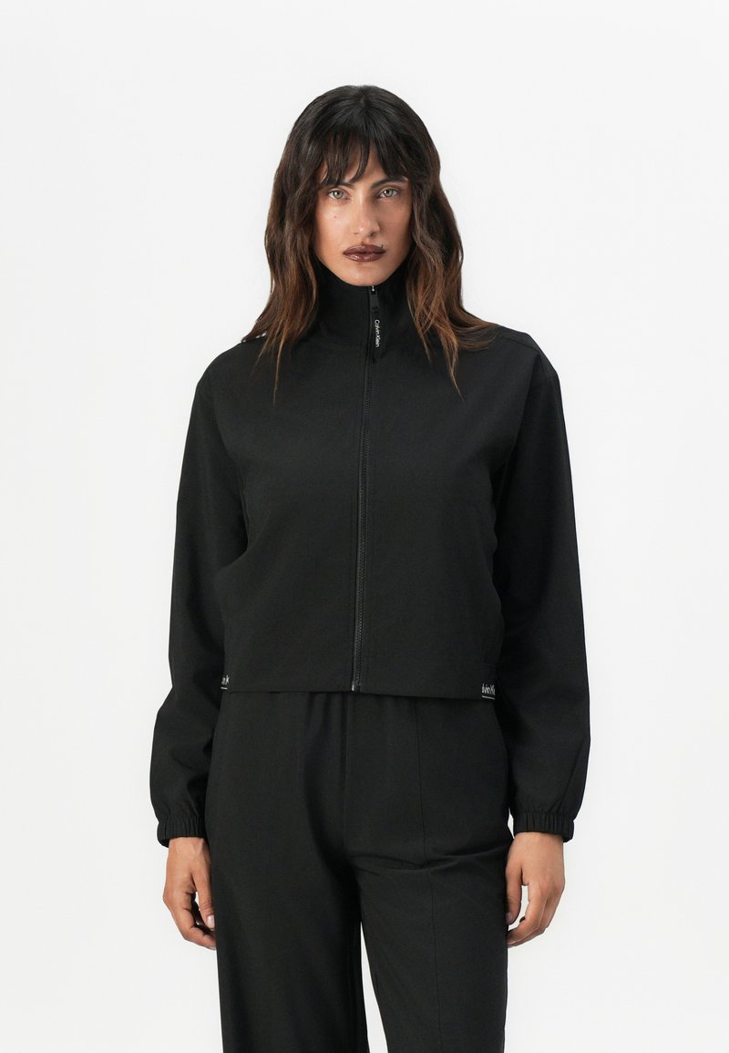 Zwarte cropped jack met een hoge kraag, ritssluiting aan de voorkant en elastische manchetten. Gemaakt van gladde stof, met subtiele branding aan de zoom.