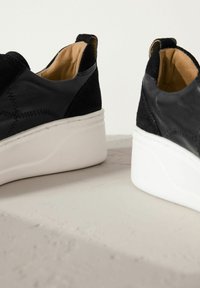 Black leather and suede slip-on shoes with a white platform sole. Features stitching details and a soft, tan inner lining.