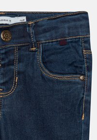 Jeans de mezclilla azul oscuro con costuras amarillas contrastantes, un bolsillo frontal con un pequeño bolsillo para monedas y un botón de marca.