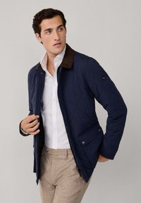 Marineblau gesteppte Jacke mit braunem Cordkragen, vorderen Taschen und silbernem Zubehör, getragen über einem weißen Hemd und hellbraunen Hosen.
