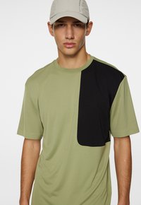 Oliivinvihreä lyhythihainen T-paita, jossa on musta paikka vasemmalla puolella. Paita on valmistettu sileästä kankaasta ja siinä on pyöreä kaula-aukko sekä rento istuvuus.