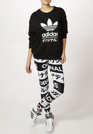 Henkilö, joka pitää päällään mustaa Adidas-hupparia ja musta-valkoisia painettuja legginssejä, pukeutuneena Adidas-tennareihin, seisoo yksi jalka ristissä.