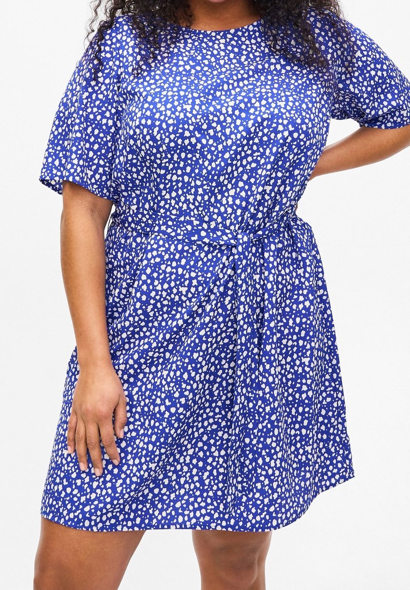 Femme portant une robe bleue à motif de points blancs irréguliers et manches courtes, une main sur la hanche et l'autre détendue le long du corps.