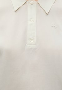 Λευκό πόλο με γιακά, τρία κουμπιά και μικρό κεντημένο λογότυπο κροκοδείλου στο στήθος.