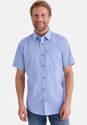 Lächelnder Mann, der ein hellblaues, kurzärmeliges Hemd mit Knöpfen und Brusttasche trägt, dazu blaue Jeans, stehend vor einem weißen Hintergrund.