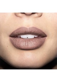 Rouge à lèvres nude mat sur des lèvres légèrement texturées, avec une forme bien définie. Dents blanches visibles entre des lèvres closes. Fond de peau lisse.