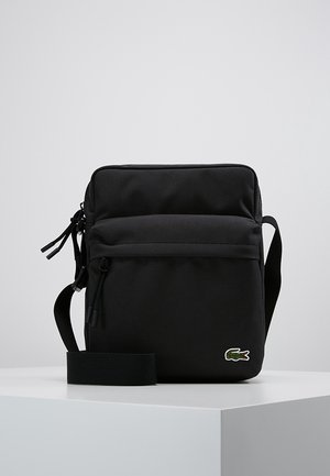 Černá kabelka přes rameno s nastavitelným popruhem, přední kapsou na zip a malým zeleným logem krokodýla vpravo dole.