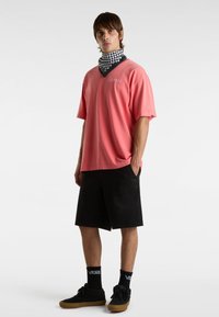 Rosa Kurzarm-T-Shirt mit lockerem Schnitt, kombiniert mit schwarzen Shorts. Accessoiert mit einem schwarz-weißen karierten Bandana und schwarzen Turnschuhen.