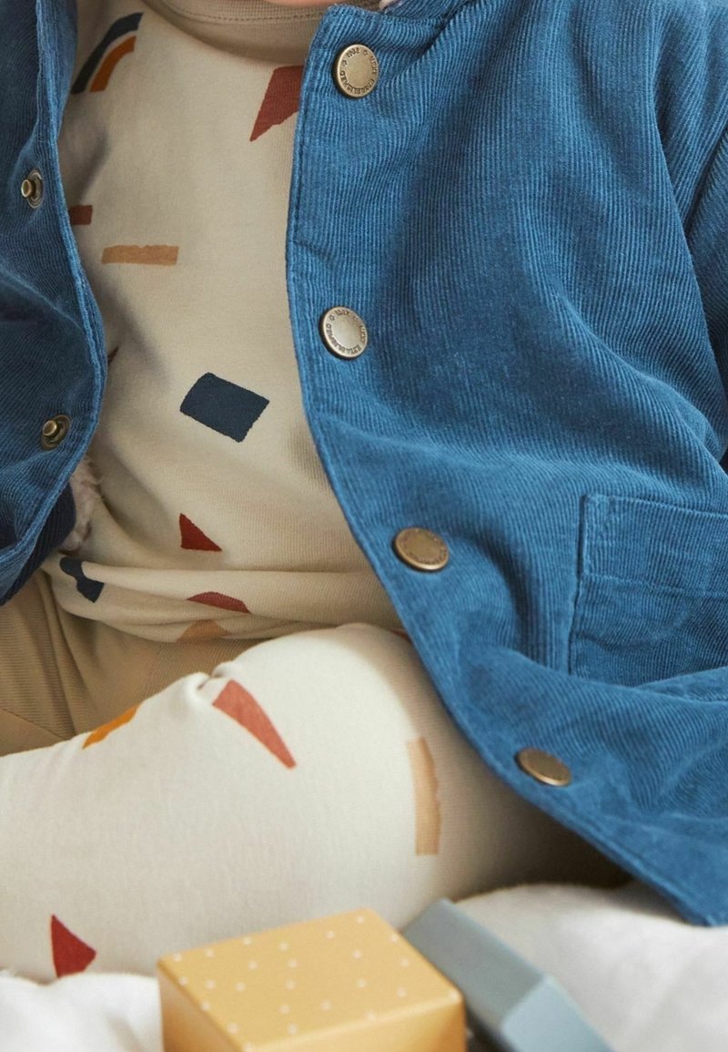 Un tout-petit portant une veste en velours côtelé bleue et une tenue crème ornée de formes géométriques colorées, assis près de blocs jouets jaunes et gris.