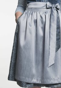 Lichtblauwe schürze met een gladde textuur en een strikband in de taille, gelaagd over een donkerder, geweven stof, met zakken en een relaxte pasvorm.