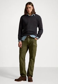 Σκούρο πλεκτό πουλόβερ, ανοιχτό μπλε πουκάμισο με γιακά από κάτω, χακί παντελόνι cargo με τσέπες, καφέ παπούτσια με κορδόνια, σε στάση όρθια.