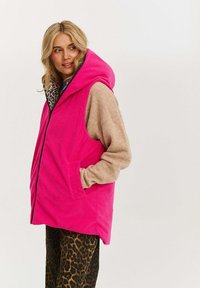 Fuchsiový kordurový vest s kapucí, který má podšívku s leopardím potiskem a béžové rukávy. Obsahuje boční kapsy a přední zipové zapínání.