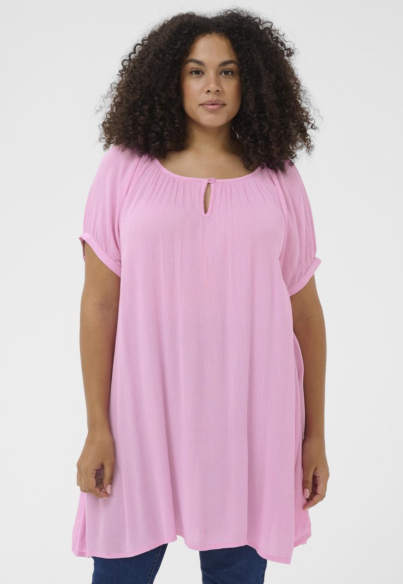 Žena s kučeravými vlasmi, oblečená v voľnej svetloružovej tunike s krátkym rukávom cez tmavomodré džínsy na jednolkom pozadí.