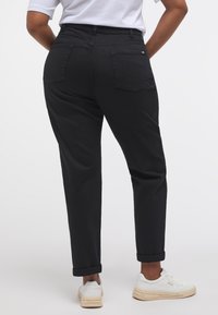 Egy személy fekete, felgöngyölt szárú nadrágot és fehér sportcipőt visel, bemutatva a hátsó nézetet és a hátsó zsebek részleteit egy sima háttér előtt.