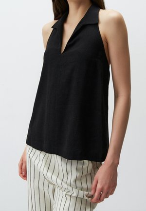Femme portant un haut noir sans manches avec col et un pantalon blanc à rayures noires verticales, tenant le tissu du pantalon d'une main.
