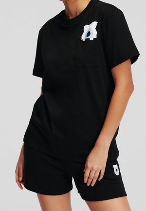 Osoba ubrana w czarną koszulkę z krótkim rękawem i szorty z białymi wzorami kreskówkowego psa na kieszeni koszulki oraz nogawce szortów.