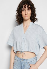 Camisa curta azul clara com mangas curtas, riscas brancas verticais, colarinho em V e detalhe de laço na barra. Fecho de botão na frente.