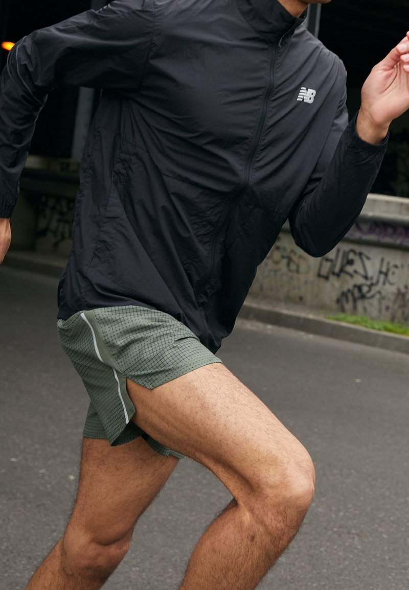 Coureur portant une veste zippée noire et un short vert, capturé en pleine foulée dans une rue urbaine avec des graffitis en arrière-plan.