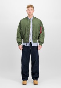 Olivengrønn bomberjakke med en glatt, blank tekstur, med glidelås foran, ribbestrikkede mansjetter og en rød labeldetalj. Kombinert med mørkeblå jeans.