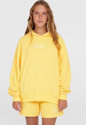 Ensemble en sweat à capuche jaune et en shorts, fabriqué dans un tissu doux. Le sweat à capuche possède une poche frontale et un texte blanc, tandis que les shorts affichent un logo assorti.