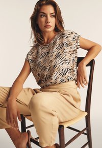 Top de estampado animal en beige con rayas negras, de mangas cortas. Pantalones color crema, de corte relajado, con textura suave, sentado en una silla de madera.