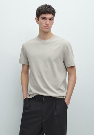 Ung mand iført en lysebeige T-shirt med korte ærmer og sorte bukser, stående med hænderne i lommerne mod en ensartet lys baggrund.