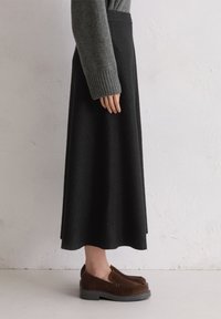 Persoon in een grijze sweater, zwarte midi rok en bruine instappers staat tegen een witte textuurwand, zij-aanzicht.