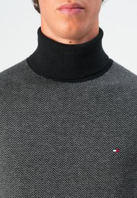 Hombre vestido con un suéter negro de cuello alto con un patrón de espiga gris y un pequeño logotipo rojo, blanco y azul en el pecho.