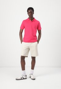 Lyserød polo med krave, parret med lyse beige shorts. Modellen har hvide og sorte sneakers på og står med hænderne i lommerne.