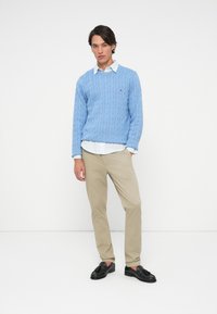 Young man wearing a light blue cable-knit sweater over a white shirt, beige pants, and black tassel loafers, standing against a white background.