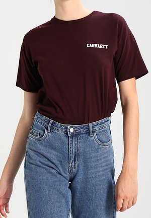 Persoon met een wijnrood Carhartt T-shirt dat in een hooggetailleerde blauwe spijkerbroek is gestopt, staand tegen een effen achtergrond.