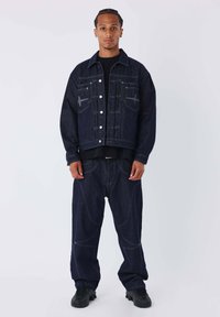 Giacca di denim scuro con cuciture e tasche visibili, abbinata a jeans scuri e larghi coordinati. Scarpe da ginnastica nere completano il look.