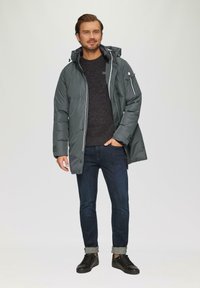 Grau gefütterte Jacke mit Kapuze, Reißverschluss vorne und seitlichen Taschen; getragen über einem dunklen Pullover und dunklen Jeans mit aufgerollten Säumen. Schwarze Sneaker.