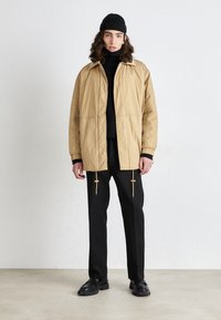 Lichtbruine gewatteerde jas met een kraag, gecombineerd met een zwarte turtleneck en zwarte broek. Voorzien van trekkoorden en zwarte schoeisel.