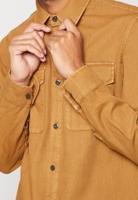 Brązowa koszula z długim rękawem wykonana z miękkiego materiału, z kołnierzykiem na guziki, dwoma kieszeniami na piersi oraz metalowymi guzikami na mankietach i z przodu.