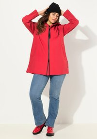 Cappotto impermeabile rosso con cappuccio, zip nera e coulisse. Due tasche frontali, dettagli con bottoni sull'orlo, abbinato a jeans azzurri e scarpe rosse.